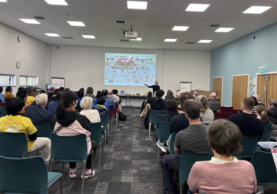 Audience seated in a lecture room watching a speaker presenting at the front with slides projected on a screen.