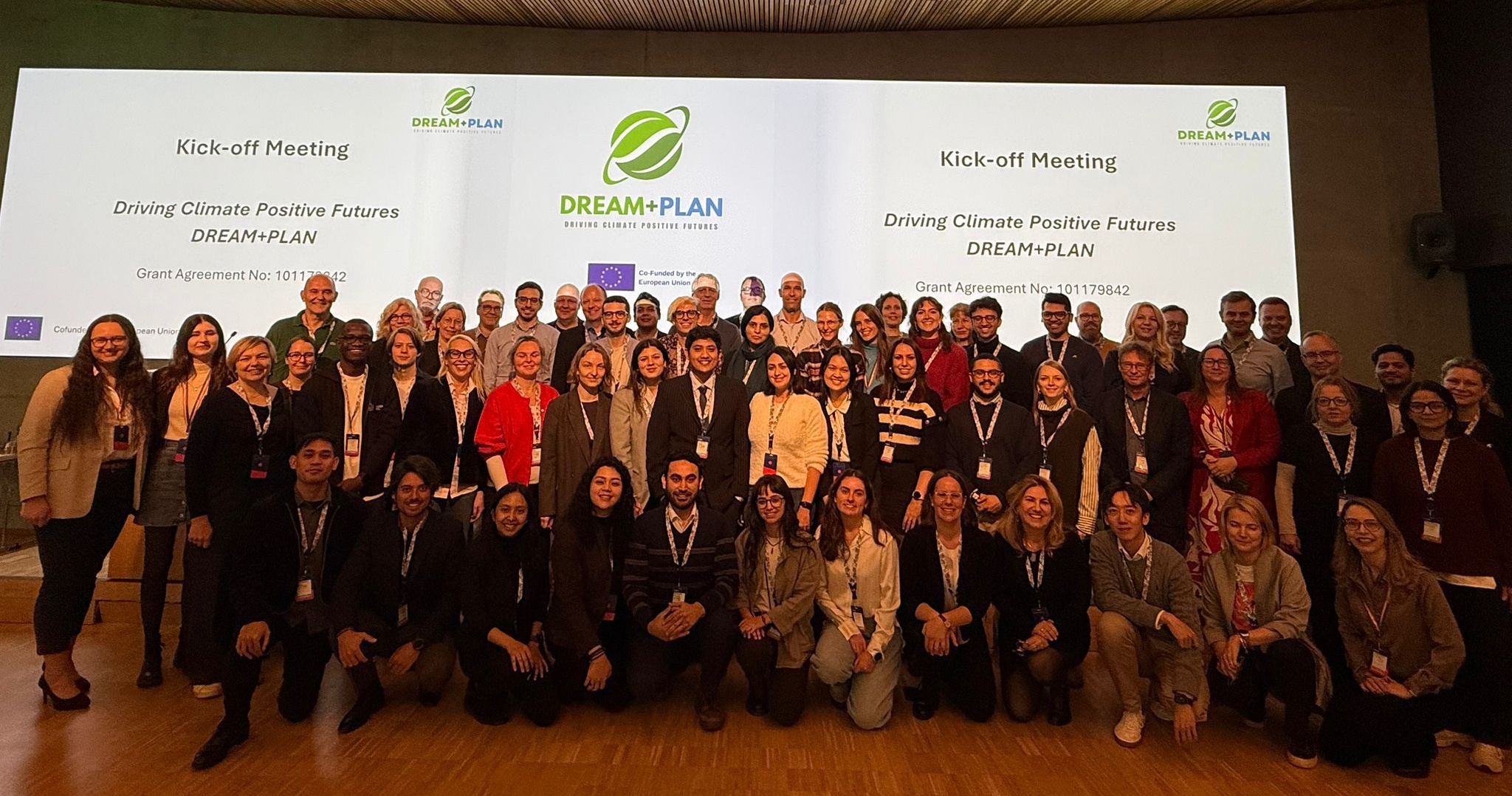 A large group of professionals posing for a photo on a stage during a kick-off meeting. Behind them, a wide screen displays the "DREAM+PLAN" logo with a green leaf icon, the slogan "Driving Climate Positive Futures," and the European Union flag indicating co-funding. The participants are arranged in several rows, with the front row kneeling and the back rows standing, all smiling towards the camera.