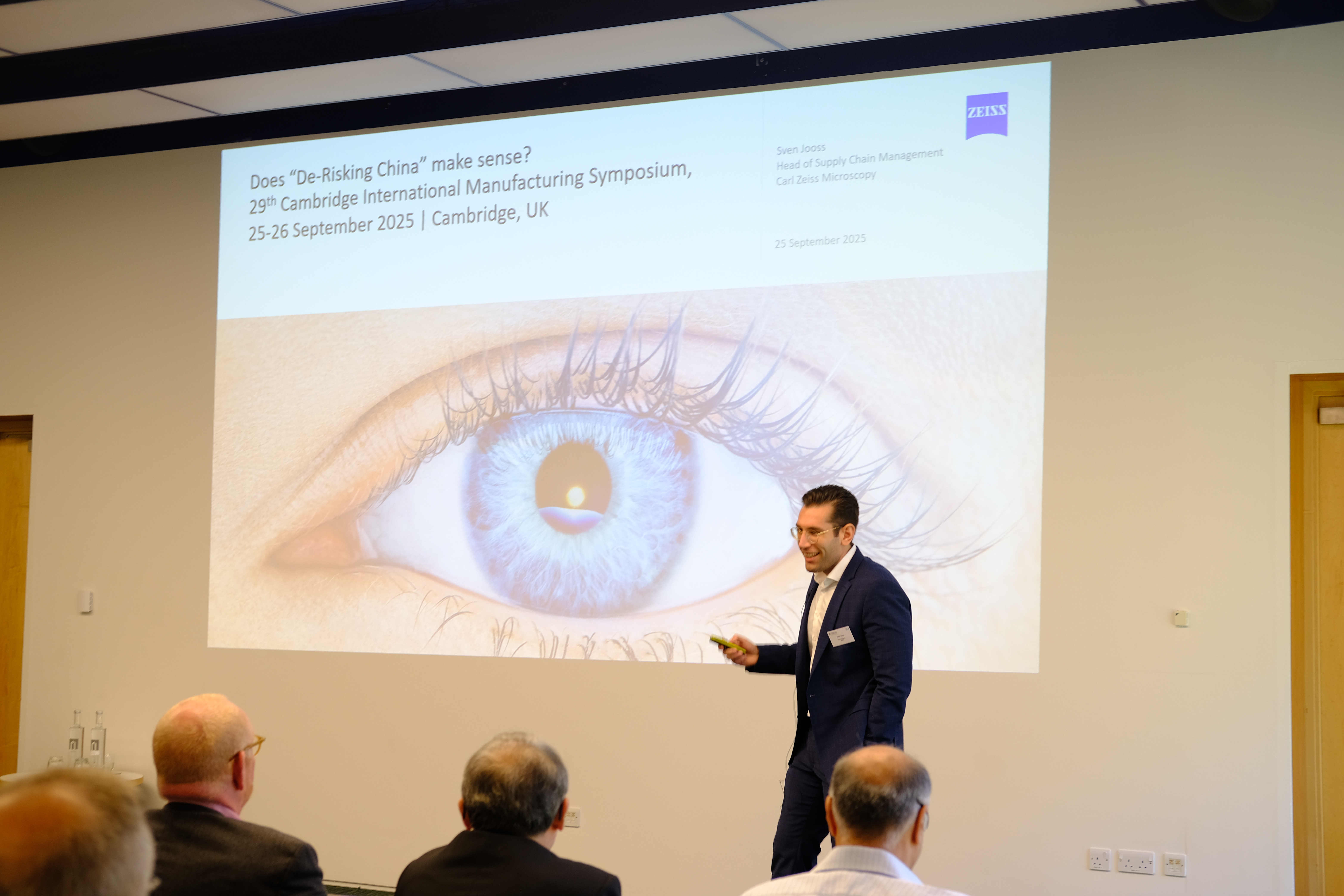A man in a suit gives a presentation at the Cambridge International Manufacturing Symposium. He stands in front of a large screen that displays a slide with the title 'Does 