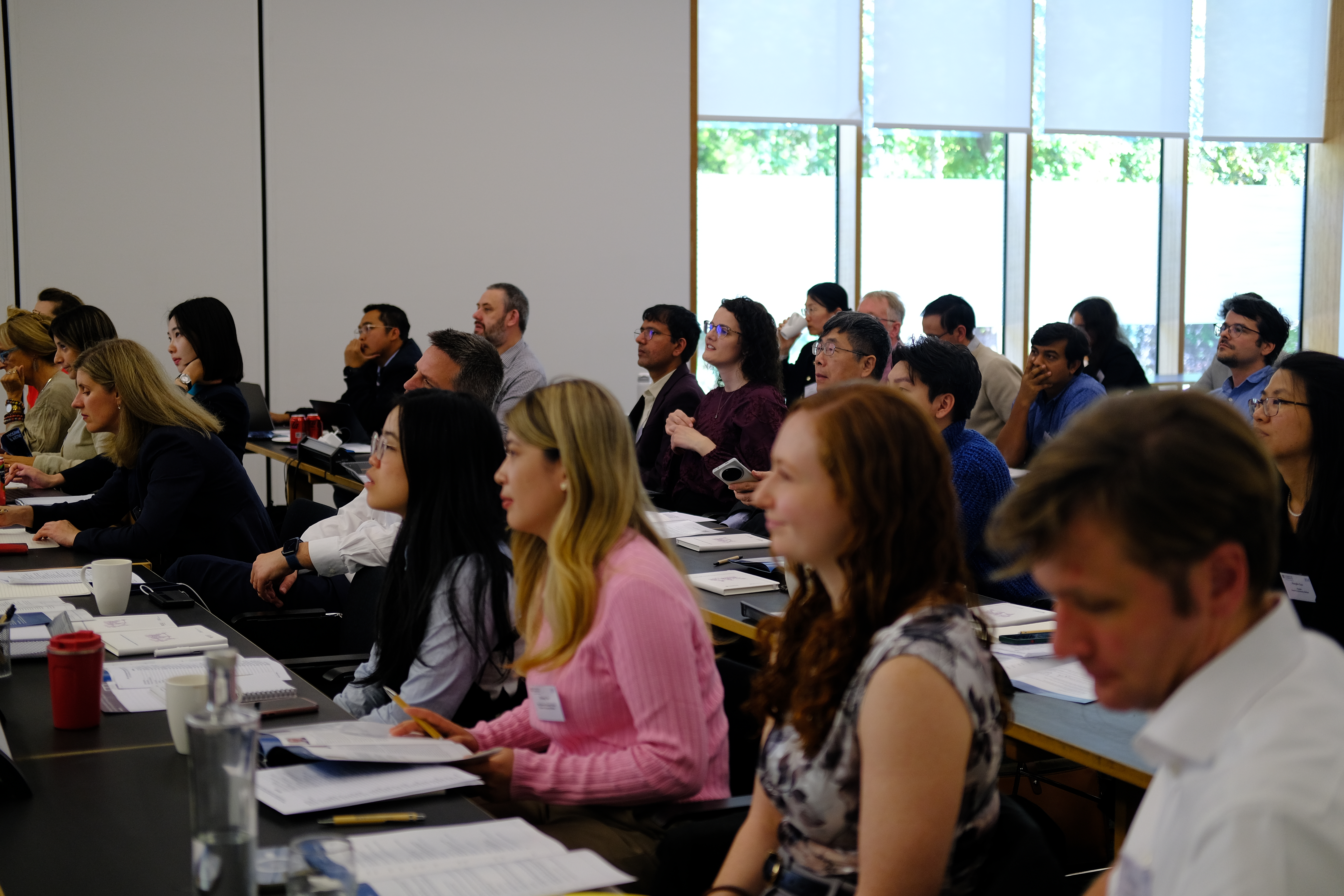 A side-view shot of an attentive audience sitting at long tables in a brightly lit lecture hall. They are listening to a presentation, with papers, laptops, and water glasses in front of them.