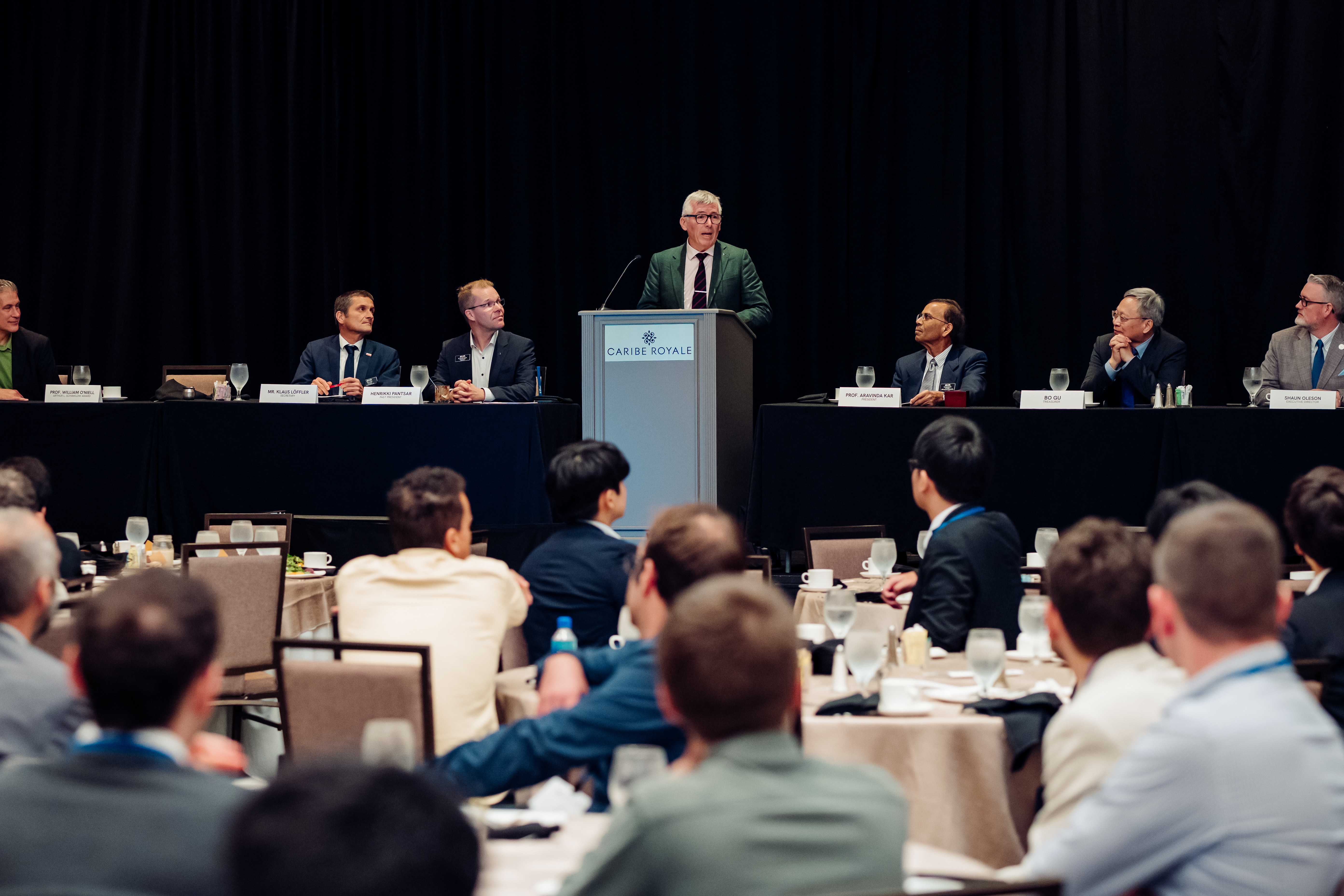 A man with grey hair and glasses, wearing a suit jacket, stands at a podium giving a speech to a large audience. On the stage behind him, seven other men in business attire are seated at a long table. The audience is seated at round tables in a conference hall, viewed from behind.